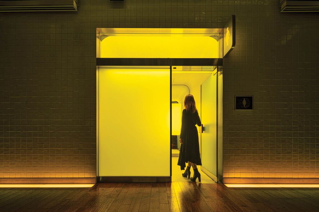 a woman opens the door to a yellow hued public toilet in Japan